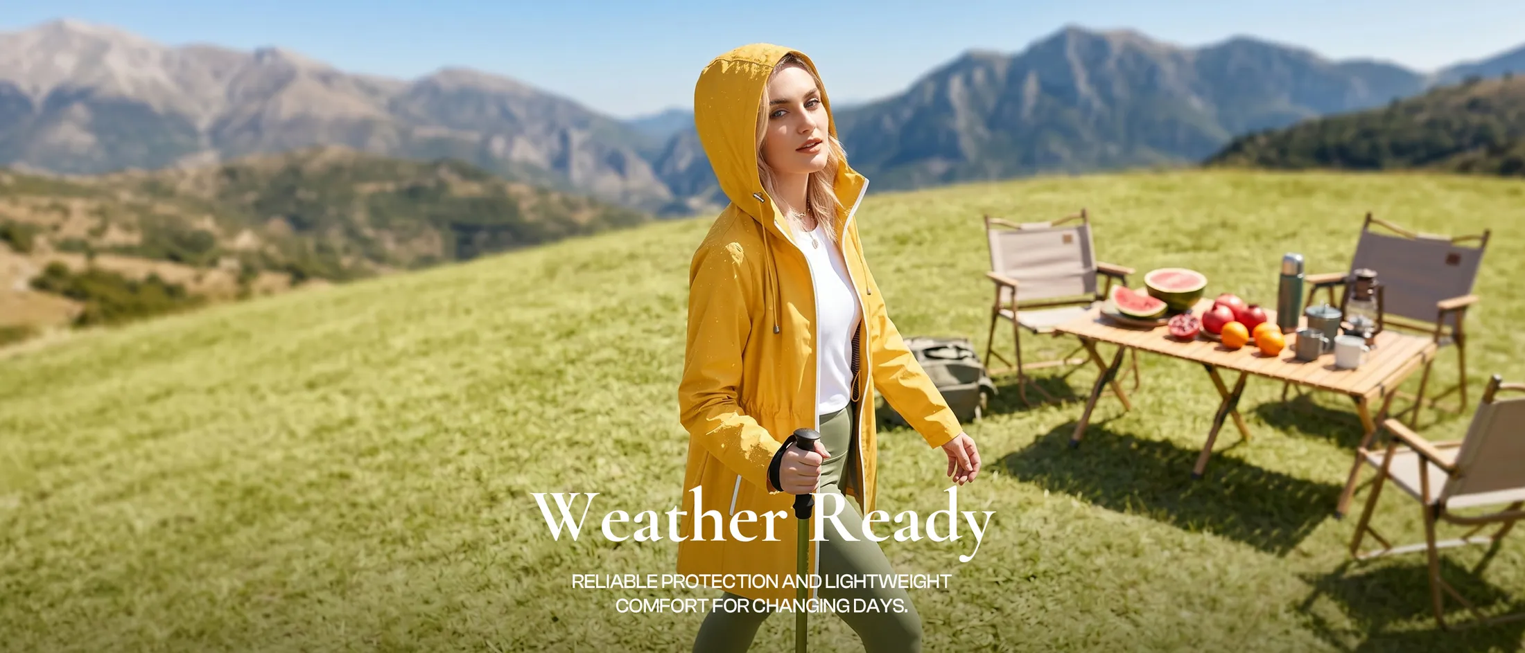 A woman in a yellow hooded shell on a mountain meadow beside a folding table